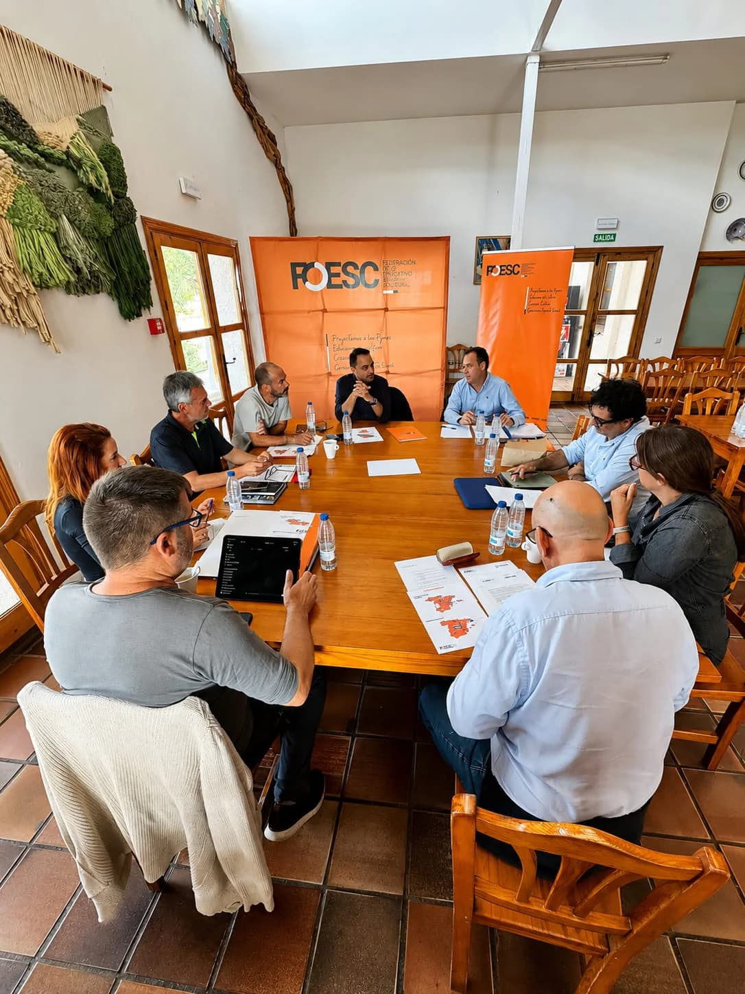 Un grup de persones es reuneix al voltant d’una taula amb suports de FOESC al fons.